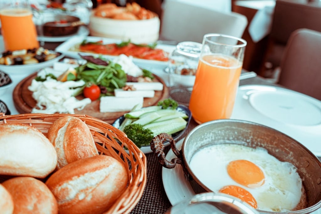 Table remplie de nourriture : oeufs au plat, pain, légumes, jus de fruits...