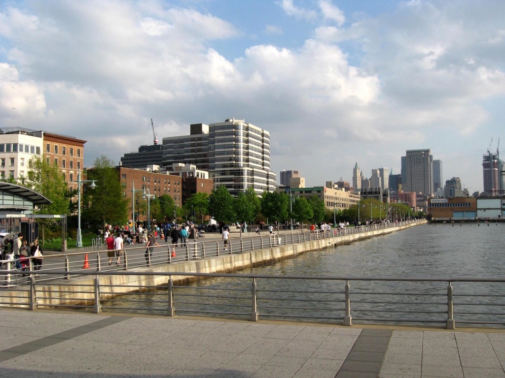 View of the Hudson River Park. There are people walking along the river and buildings in the background.